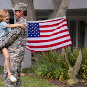 Caucasian soldier father carrying son in garden with american flag hanging outside house. soldier returning home to family.