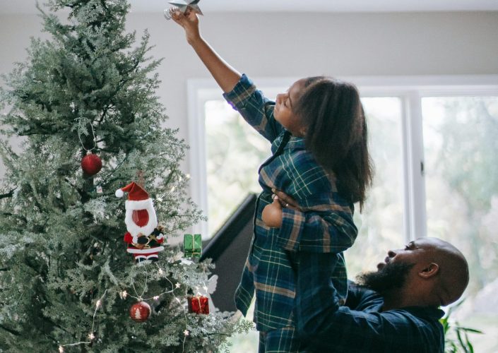 Father lifts daughter to place star on Christmas tree, celebrating holiday spirit indoors.
